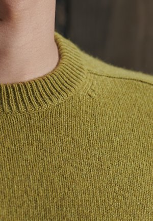 Zbliżenie osoby noszącej prążkowany, oliwkowozielony dziergany sweter z okrągłym dekoltem na rozmytym, ciemnym tle.