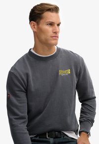 Joven con unsuéter oscuro de Superdry, camiseta blanca, pantalones vaqueros azules, un cinturón negro y un reloj en la muñeca, mirando a su derecha.