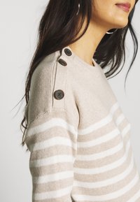 Krémovo-bílý pruhovaný svetr s kulatým výstřihem, který má hnědé knoflíkové detaily na rameni a měkké, strukturované pletení.