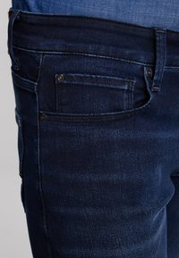 Tumši zili džinsi tuvu skatījumā, rādot priekšējo kabatu ar monētu kabatu un redzamām kniedēm un šuvju detaļām.