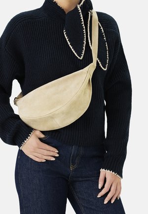 Beige suède schoudertas met een gebogen vorm, verstelbare band en gladde textuur, gedragen over een zwarte geribbelde trui en donkere jeans.