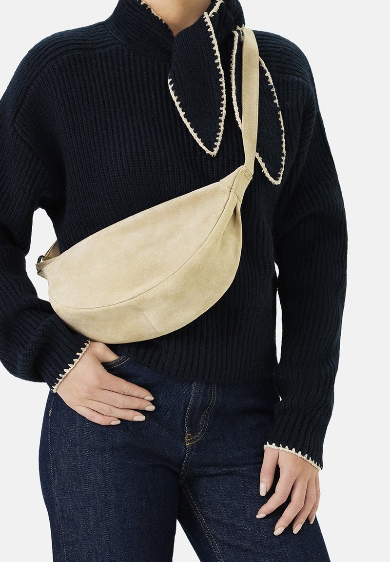 Beige suède schoudertas met een gebogen vorm, verstelbare band en gladde textuur, gedragen over een zwarte geribbelde trui en donkere jeans.