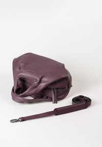 Borsa in pelle viola con doppia cerniera e tracolla staccabile, arrotolata accanto a essa su uno sfondo bianco.