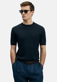 Marineblauwe geribbelde gebreide sweater met korte mouwen en een ronde halslijn, gecombineerd met lichtblauwe geplooide broek en een bruine leren riem; model draagt een zonnebril.