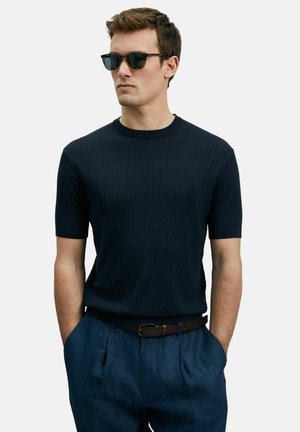 Pull en maille côtelée marine à manches courtes avec un col rond, associé à un pantalon plissé bleu clair et une ceinture en cuir brun ; le modèle porte des lunettes de soleil.