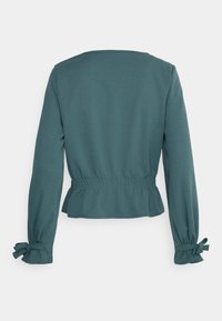 Teal long-sleeve top with a relaxed fit, smocked waist, and bow ties at the sleeve cuffs. Smooth texture, simple round neckline.