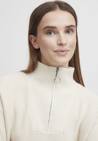 Kreemivärvi koe tõmblukuga pullover kõrge kraega. Omab soonikstruktuuri ja esitaskut. Hõbedane tõmblukk.