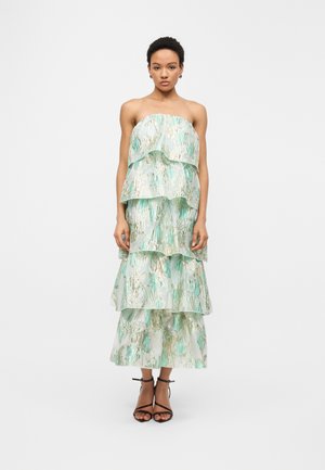 Kobieta ubrana w sukienkę bez ramiączek z falbanami, w miętowo-złoty kwiatowy wzór, zestawioną z czarnymi sandałami na wysokim obcasie z paskami.