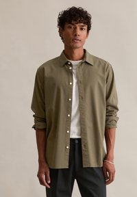 Oliwkowa koszula zapinana na guziki z kołnierzykiem, podwiniętymi rękawami i białymi guzikami, noszona na białym t-shircie, zestawiona z ciemnymi spodniami.