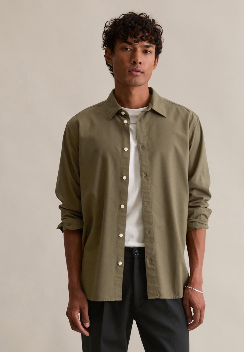 Oliwkowa koszula zapinana na guziki z kołnierzykiem, podwiniętymi rękawami i białymi guzikami, noszona na białym t-shircie, zestawiona z ciemnymi spodniami.