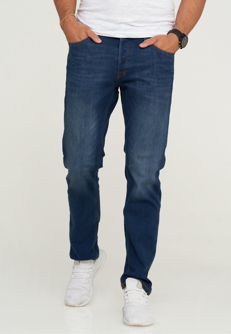 Mörkblå denimjeans med slim fit, med en urtvättad look, femficksdesign och kontrasterande orange sömmar, bärs med vita sneakers.