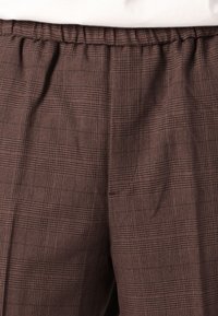 Braune karierte Hose mit elastischem Bund, aus einem strukturierten Stoff, der über das ganze Material ein feines Karomuster zeigt.