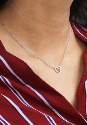Zilveren hartvormige ketting met een kleiner hartje binnenin, voorzien van een ketting en sprankelende accenten, gedragen op een bordeaux gestreepte blouse.