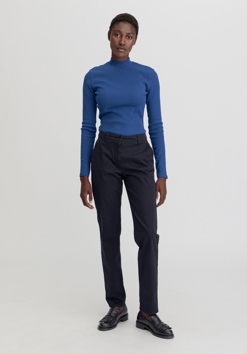 Niebieska dopasowana bluzka z długim rękawem zestawiona z wąskimi czarnymi spodniami i czarnymi loafersami. Gładka faktura, prosty design oraz neutralne tło.