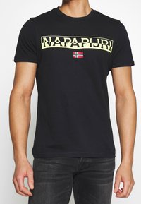 Camiseta de algodón negra con mangas cortas, que presenta un amplio logo amarillo de "NAPAPIJRI" y un pequeño gráfico de la bandera noruega en el centro.