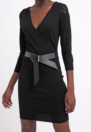 Kvinde iført sort omslagskjole med trekvartlange ærmer, blonder ved skuldrene, og bred sort bælte med guldlås ved taljen.