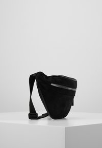 Borsa nera in suede a forma triangolare, con zip sulla parte superiore, tracolla regolabile e superficie testurizzata. Design minimalista.