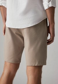 Homme portant un short beige et une chemise blanche à manches longues retroussées, vu de dos au niveau de la cuisse.