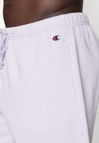Pantalons de jogging lavande clair en tissu doux, dotés d'une taille élastique, de poches latérales et d'un petit patch logo sur la cuisse.
