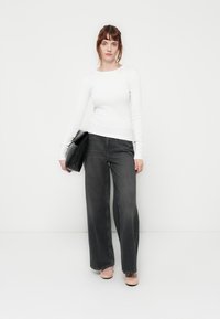 White long-sleeve fitted top paired with high-waisted, wide-leg dark gray jeans. Model holds a black clutch and wears light-colored shoes.