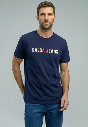 Hombre con cabello corto y oscuro y barba, con una camiseta azul marino con el texto "SALSA JEANS" y vaqueros azules, de pie frente a un fondo liso gris claro.