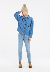 Veste en polar bleue avec deux poches à l'avant et des boutons marron, associée à un jean en denim clair et un bonnet en tricot beige avec un pompon sur le dessus.