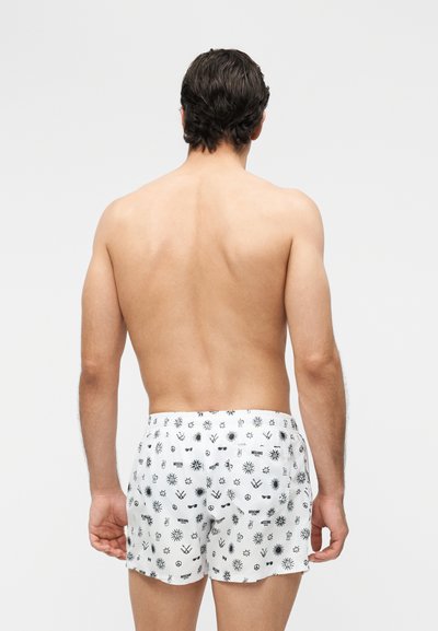 Mann sett bakfra iført hvite shorts med sorte symboler som sol, fredstegn og andre små på en ensfarget lys grå bakgrunn.