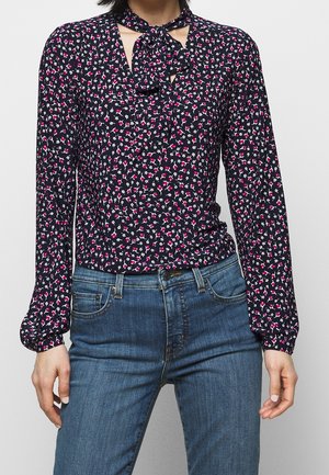 Femme portant un chemisier noir à fleurs à manches longues avec un détail nœud au col, rentré dans un jean bleu.