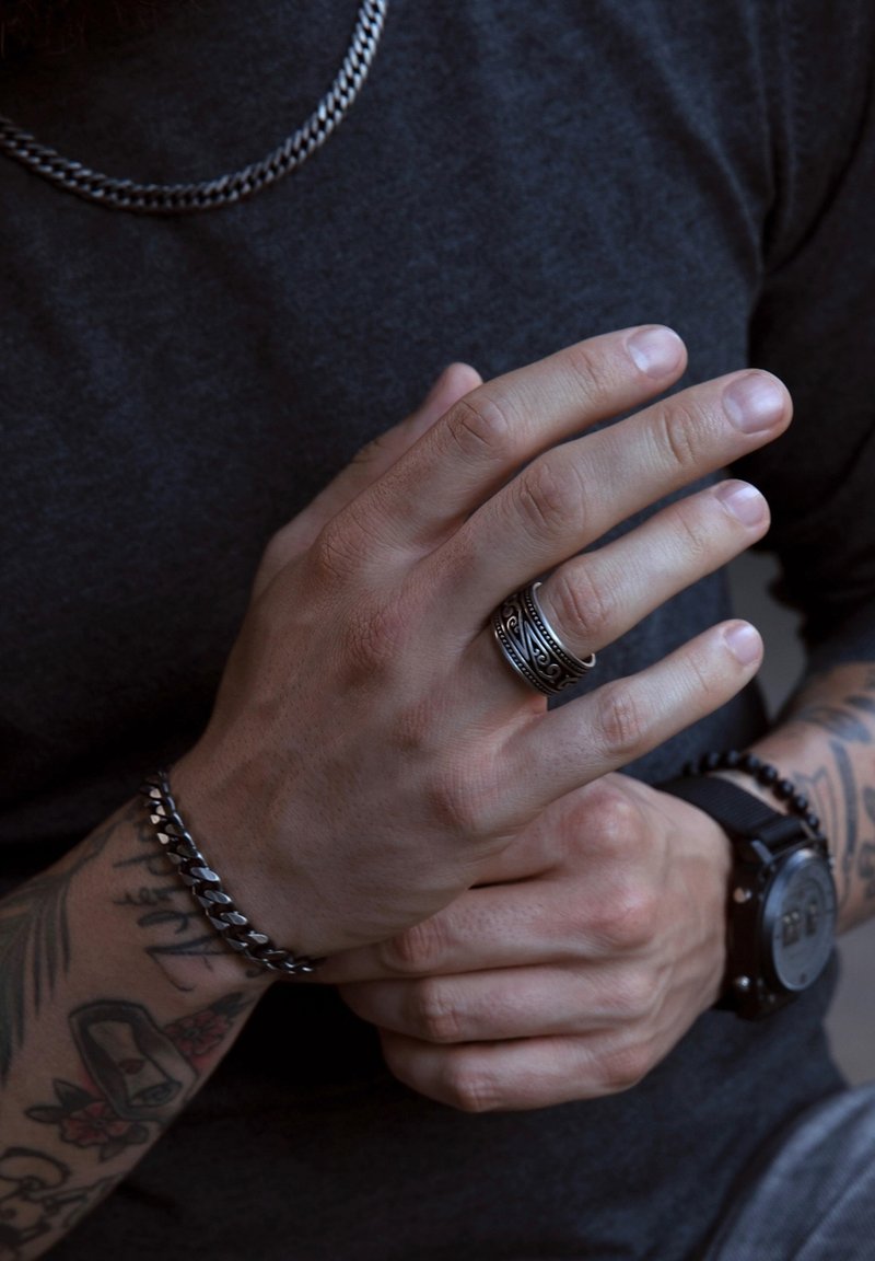 Zilveren ketting en armband op een hand met een zwarte ring met ingewikkelde patronen; huidtint en tatoeages zichtbaar.