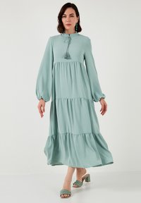 Long mint-green tiered dress with a high neckline, gathered sleeves, and tassel detail. Paired with matching sandals featuring ruffled accents.