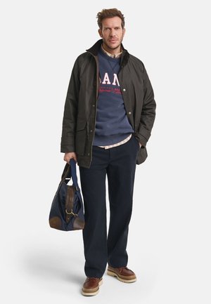 Donkerbruine jas, marineblauwe hoodie met rode letters, lichtgeruit shirt, donkere broek, bruine schoenen en een marineblauwe tas met bruine accenten.