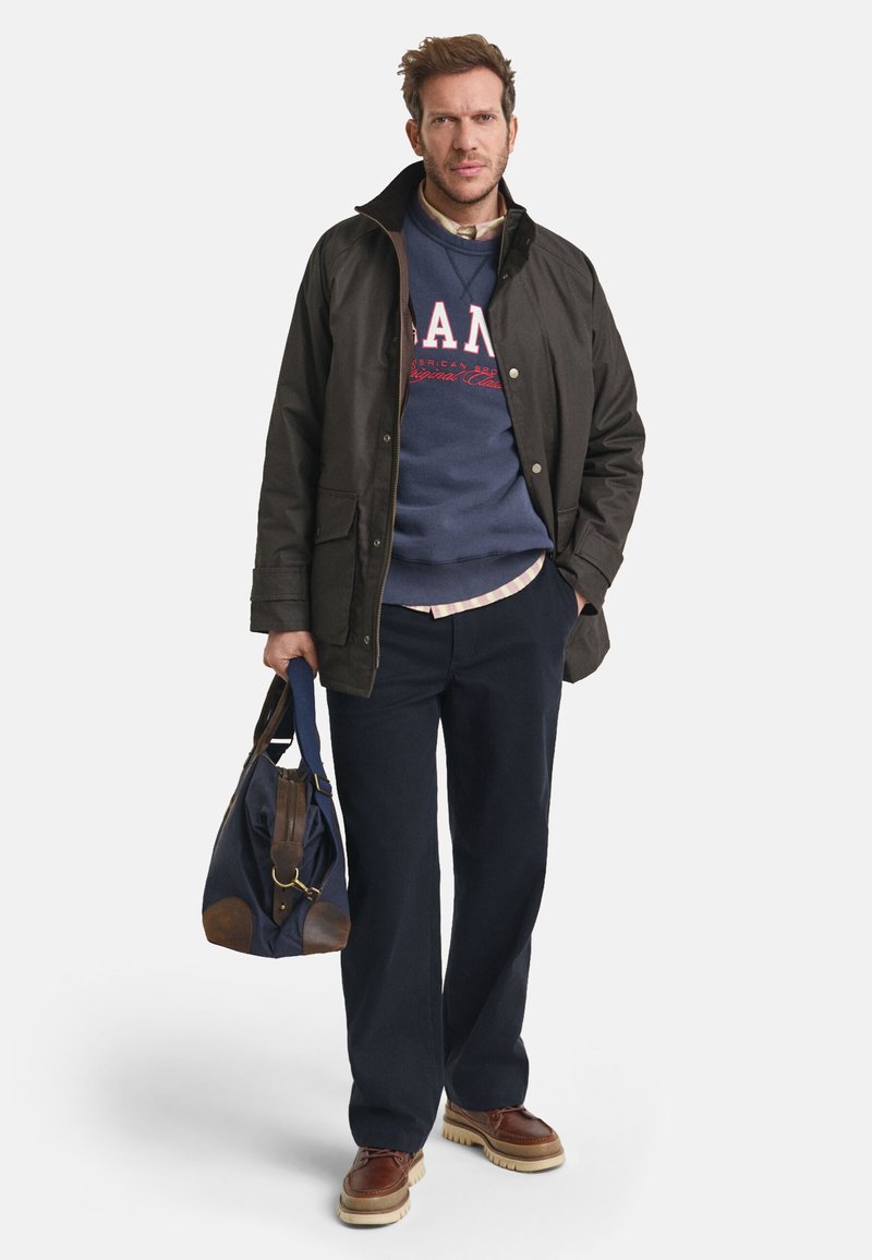 Donkerbruine jas, marineblauwe hoodie met rode letters, lichtgeruit shirt, donkere broek, bruine schoenen en een marineblauwe tas met bruine accenten.