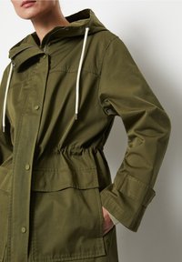 Olivgrön parka-jacka med dragsko i huvan, elastisk midja, framfickor och tryckknappsstängning. Slät tyg med matt finish.