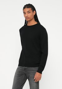 Zwarte gebreide trui met een ronde halslijn, lange mouwen en een strakke tailleband. Gecombineerd met grijze denim jeans.