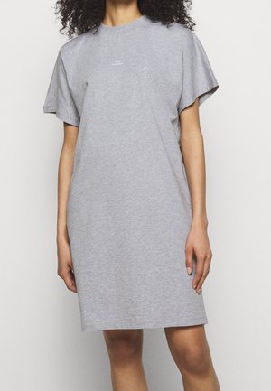 Vrouw draagt een losse, korte mouw grijze t-shirtjurk met kleine witte tekst op de borst, staand tegen een effen lichte achtergrond.