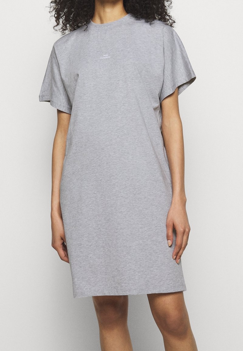 Femme portant une robe t-shirt grise ample à manches courtes avec un petit texte blanc sur la poitrine, debout devant un fond clair uni.