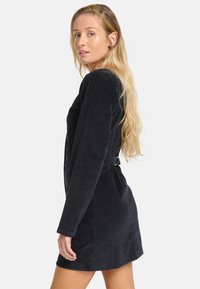 Schwarzes Cordkleid mit langen Ärmeln, tailliertem Schnitt und einer Schnalle auf der Rückseite. Glatte Textur, knielange Länge und strukturierte Form.