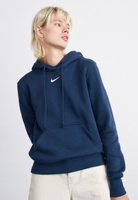 Tamsiai mėlyna džemperis su kengūros kišeniu priekyje, su raišteliu reguliuojama kapučino, ir baltu Nike logotipu ant krūtinės. Minkšta medžiaga su gaubteliuose ir apykaklėje.
