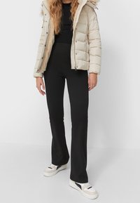 Polstret lys beige jakke med hætte, sort tætsiddende bluse, sorte flagrende bukser og hvide sneakers med sorte detaljer. Glat tekstur hele vejen igennem.