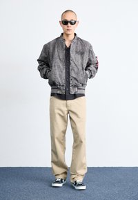 Gray bomber jacket with a textured finish, buttoned black shirt, beige trousers, and black sneakers with white soles. Model stands with hands in pockets.