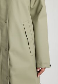 Water-resistant, light olive green coat with a soft texture, snap buttons, and a side pocket. Features a straight cut and stitched seams.