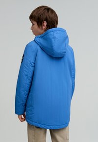 Chaqueta acolchada azul con capucha, de tejido texturizado y detalle en parche negro en la manga. Presenta un dobladillo trasero más largo.