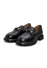 Une paire de mocassins en cuir noir avec des semelles épaisses et des détails en métal doré en forme de mors sur le dessus, présentée sur fond blanc.