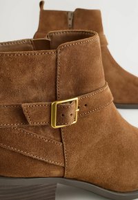 Stivali ankle in suede marrone con punta rotonda, dotati di un accento di fibbia dorata e cerniera laterale. Superficie testurizzata con dettagli di cucitura.