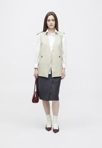 Femme aux longs cheveux bruns portant une chemise blanche, un gilet beige, une jupe foncée, des chaussettes blanches, des chaussures brunes, et tenant un sac à main marron.