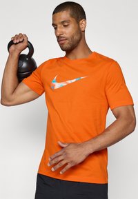 Oranje katoenen T-shirt met een blauw en gemêleerd Nike-logo op de borst, korte mouwen en een ronde hals. Kettlebell in de hand.