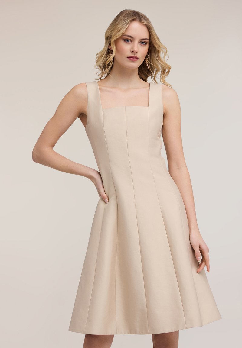 Femeie cu păr blond, purtând o rochie fără mâneci, bej, până la genunchi, cu pliuri, stând cu o mână pe șold, pe un fundal neutru.