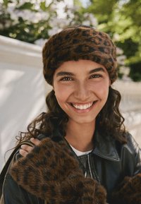 Fuskpälsmössa och muddar i brunt och svart leopardmönster, båda bärs med en svart läderjacka. Materialen har ett mjukt, strukturerat utseende.