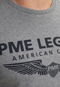 Camiseta de algodón gris con cuello redondo. Presenta un diseño gráfico en oscuro azul marino, incluyendo el texto "PME LEGEND" y acentos de escudo alado.