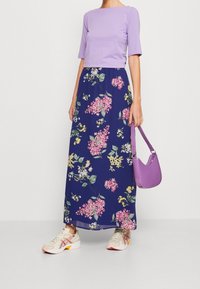 Kvinna klädd i en lila topp, marinblå blommig maxikjol, vita sneakers, som håller en lila handväska mot en vit bakgrund.